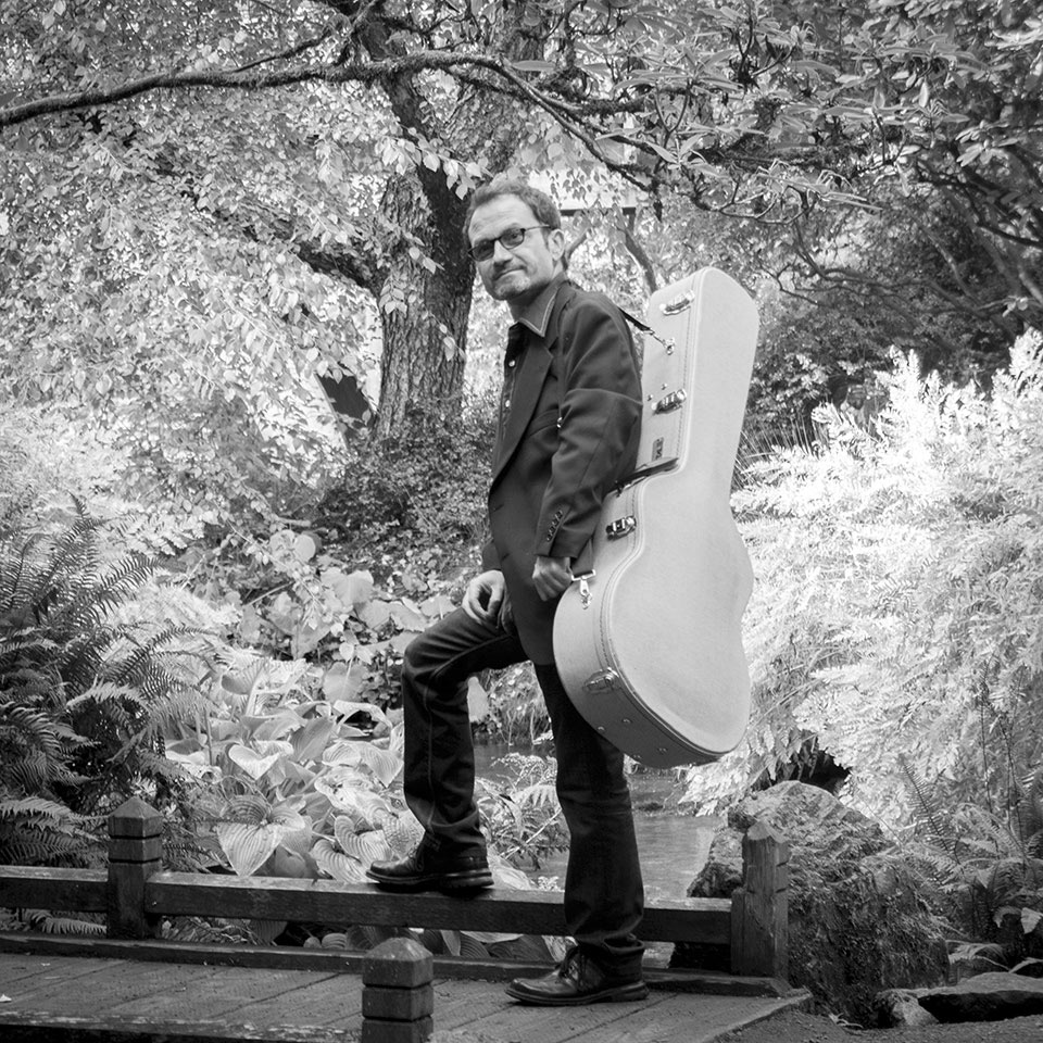 A black and white image of a man in sunglasses with a guitar case slung over his shoulder